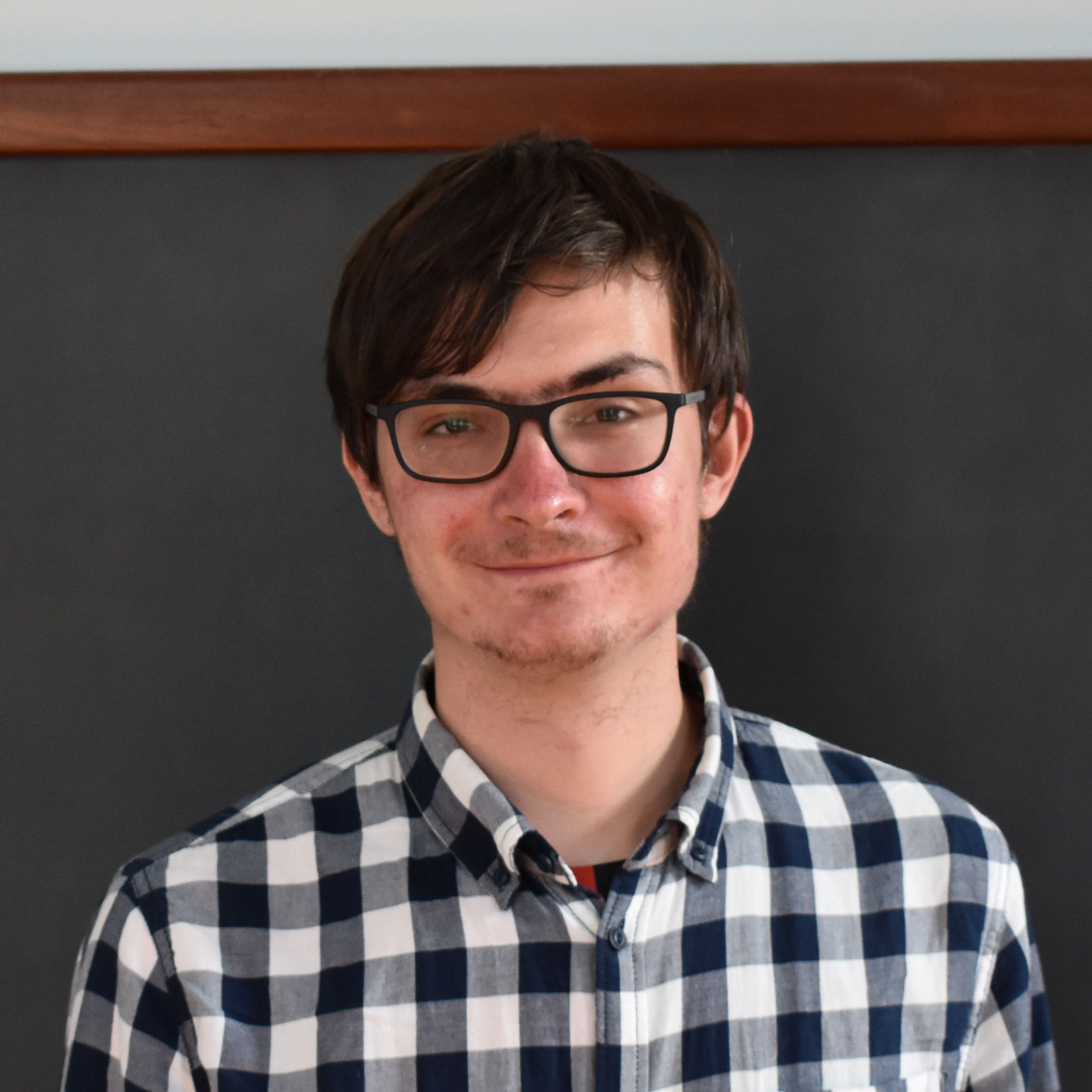 A tall young man in a checked shirt, standing in front of a blackboard, smiling.