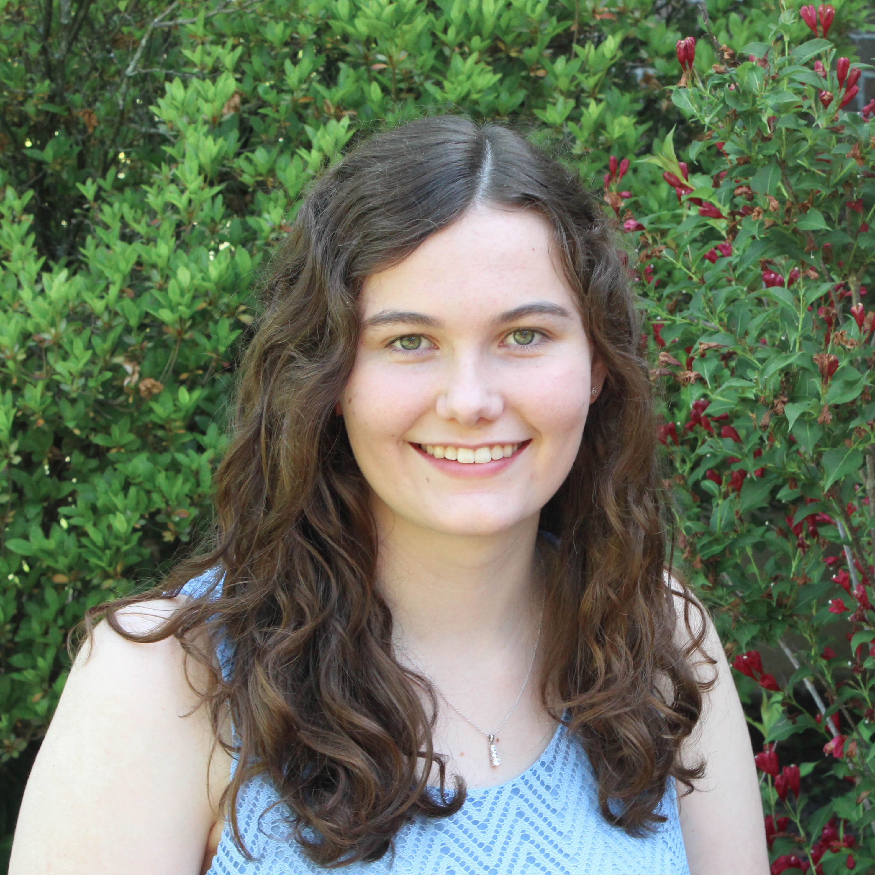 Portrait of a young woman, smiling at the camera, outside in front of a blooming hedge: Cassandra Caragine, PhD student in linguistics.