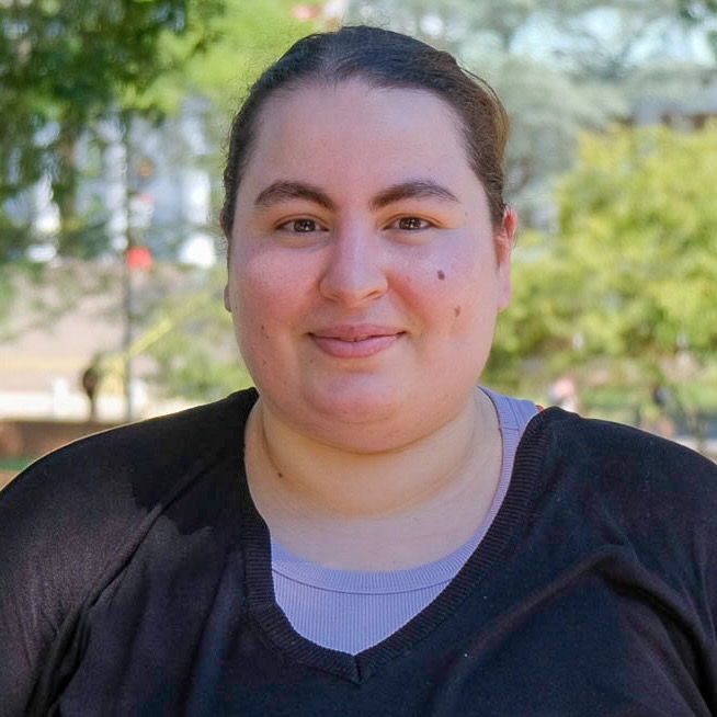 Close portrait of a young woman, Sarah Boukendour, smiling at the camera, standing outside in front of trees.