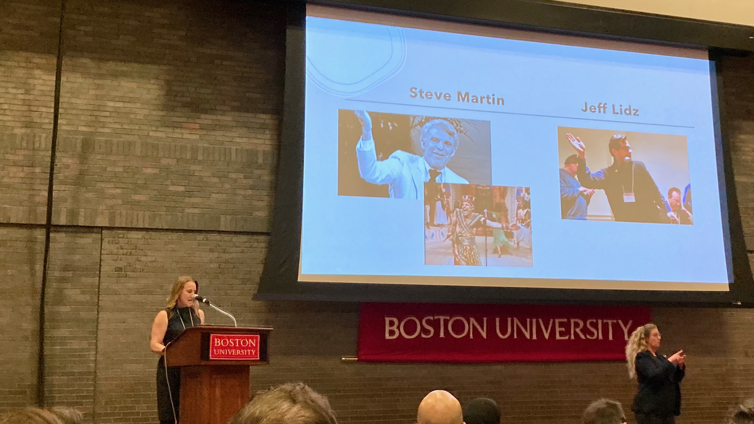 A woman at a podium in a large auditorium, with a giant screen behind her, comparing someone to Steve Martin.
