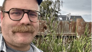 A man with a mustache standing outside in a meadow, with a basketball hoop behind him.