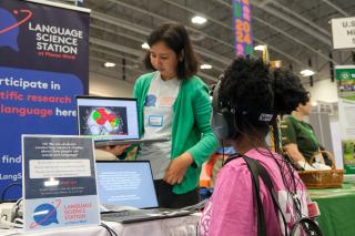 A girl holds a computer monitor showing a brain to a girl wearing an fNIRS headset